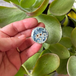 Könnte beinhalten: Ein blauer und weißer Keramikknopf mit gesprenkeltem Glasur. Der Knopf hat zwei Löcher zum Nähen.