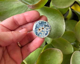 May include: A blue and white ceramic button with a speckled glaze. The button has two holes for sewing.