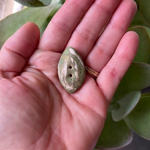 May include: A green and white ceramic button with two holes. The button is shaped like a teardrop and has a mottled glaze.