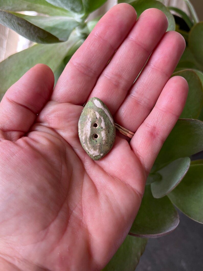 May include: A green and white ceramic button with two holes. The button is shaped like a teardrop and has a mottled glaze.