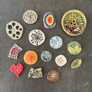 May include: An assortment of ceramic buttons in various shapes, colours, and patterns. The buttons feature designs such as a flower, a heart, a circle, and a star. The buttons are arranged on a grey surface.
