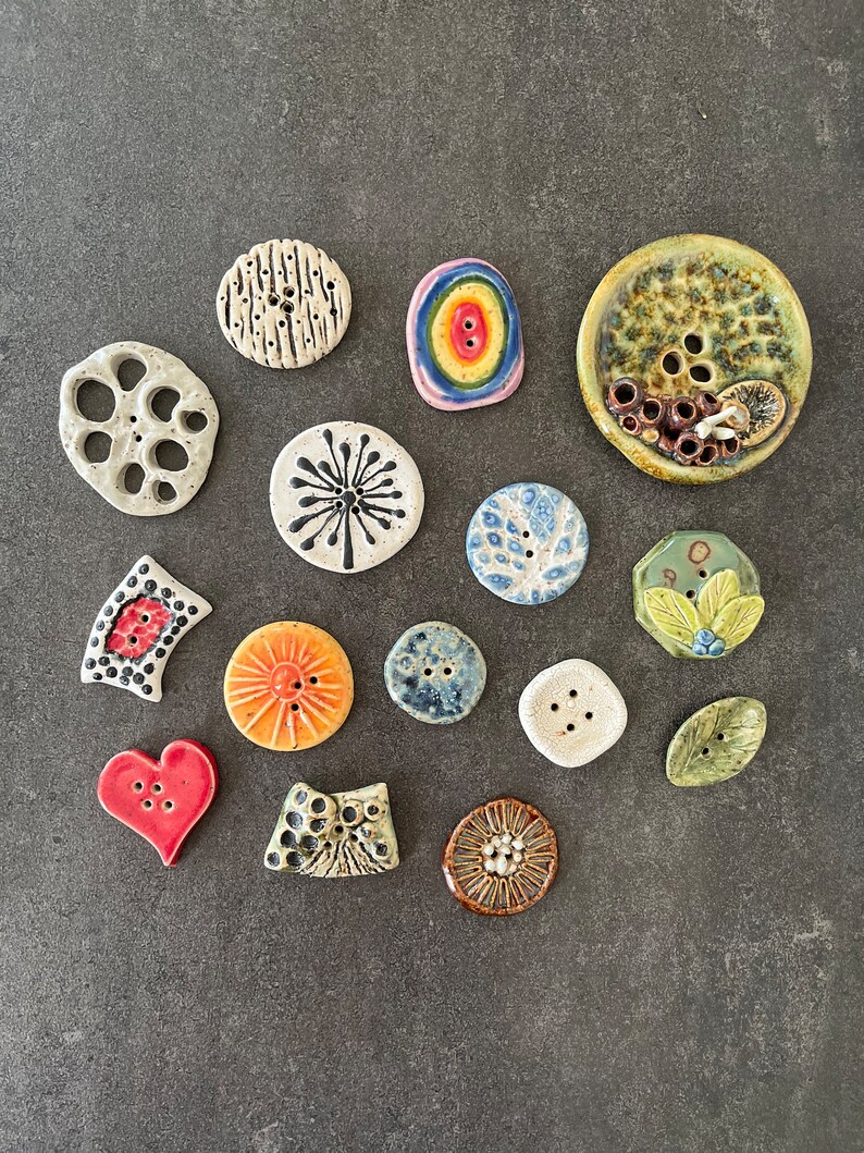 May include: An assortment of ceramic buttons in various shapes, colours, and patterns. The buttons feature designs such as a flower, a heart, a circle, and a star. The buttons are arranged on a grey surface.