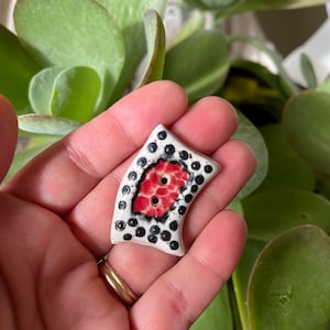 May include: A white ceramic button with a red and black design. The button has two holes and a curved shape.