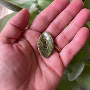 Peut inclure: Un bouton en céramique vert en forme de feuille avec deux trous pour coudre. Le bouton a un motif de feuille détaillé et un glaçage moucheté.
