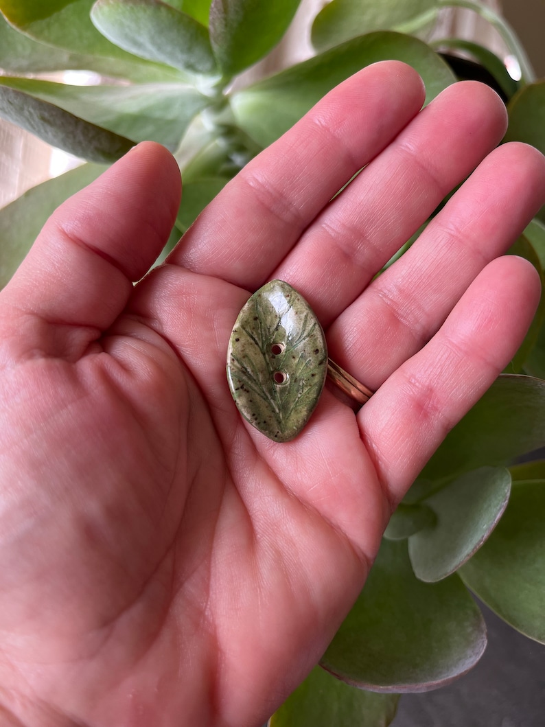 May include: A green ceramic button shaped like a leaf with two holes for sewing. The button has a detailed leaf design and a speckled glaze.