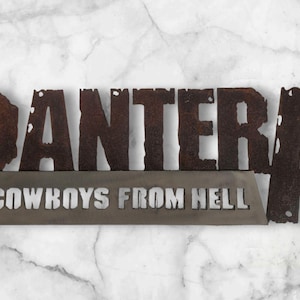 May include: Metal sign featuring the word "PANTERA" in large, distressed, brown letters above the phrase "COWBOYS FROM HELL" in a smaller, silver font. The sign is mounted on a marble-patterned surface.