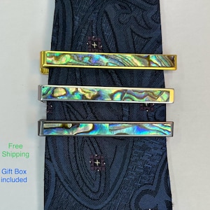 May include: Three tie clips with iridescent abalone shell inlays. The clips have gold, silver, and gunmetal finishes. The tie clips are displayed on a navy blue patterned tie.