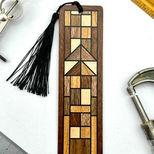 May include: A wooden bookmark with a geometric design. The bookmark features a mosaic pattern of various wood tones, including light and dark browns, and beige. A black tassel hangs from the top. The bookmark is rectangular and has a dark brown border.