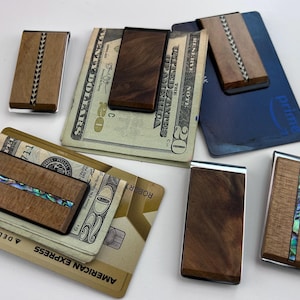 May include: A collection of wooden money clips with various designs. Some clips feature inlaid patterns of black, silver, and abalone shell. Several clips are holding cash and credit cards, showcasing their functionality and style.
