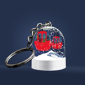 Puede incluir: Un llavero plateado con un charm de bola de nieve transparente. La bola representa un teleférico rojo con pasajeros en su interior, sobre un fondo de montaña nevada.
