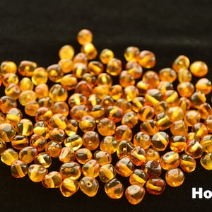 May include: A pile of honey amber beads, a type of gemstone, on a black background. The beads are round and have a smooth, polished surface. The color of the beads is a warm, golden yellow.  Honey
