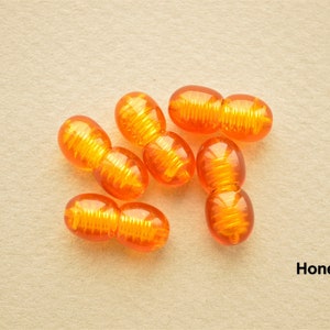 May include: A close-up of eight amber-coloured plastic beads. Each bead is made of two connected oval shapes with a textured surface. The beads are arranged in a cluster on a light brown surface. The word "Honey" is written in black text below the beads.