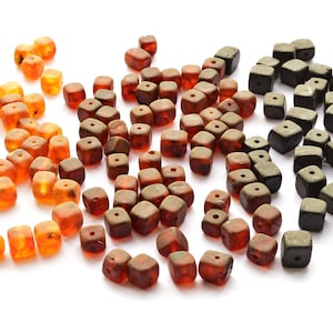 Può includere: Una collezione di perle di ambra in varie tonalità di marrone, arancio e nero. Le perle sono di forma quadrata e hanno un foro al centro per l'infilaggio.