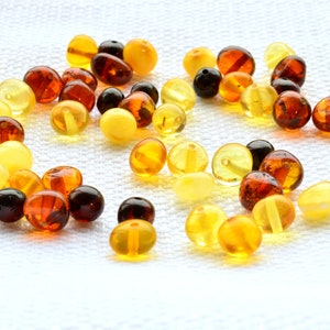 May include: A collection of amber beads in various shades of yellow, orange, and brown. The beads are round and smooth, and they are scattered on a white surface.