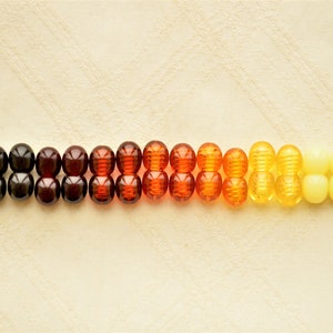 May include: A gradient of amber beads, ranging from black to yellow, arranged in a line on a beige surface.