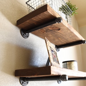 May include: Two wall-mounted shelves featuring thick, dark-stained wooden planks supported by black industrial metal pipes and circular wall flanges. The top shelf holds a black wire basket with a green plant, and the bottom shelf holds a picture frame.