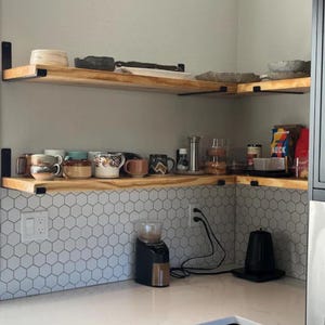 May include: Two wooden shelves with black metal brackets, mounted on a white tiled wall. The shelves are filled with mugs, dishes, and other kitchen items. There is a coffee grinder and a kettle on the counter below the shelves.