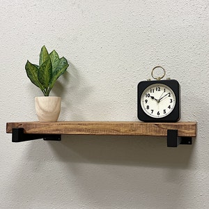 May include: A wooden shelf with black metal brackets, a potted plant, and a black alarm clock with a white face and gold accents. The clock face reads 10:10.