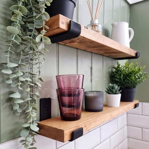 May include: Two natural wood floating shelves with matte black metal brackets mounted on a sage green wall. The top shelf holds a black pot, a reed diffuser, and a white pitcher. The bottom shelf holds two stacked purple glasses, a gray candle, and two potted plants.
