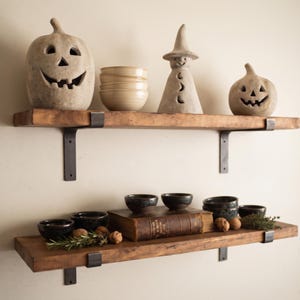 May include: Two horizontal wooden shelves with dark metal brackets mounted on a wall. The top shelf holds two carved stone jack-o'-lanterns, a stack of light-colored ceramic bowls, and a stone witch figure. The bottom shelf holds dark bowls, a book, walnuts, and herbs.