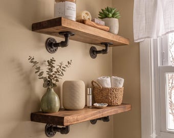 May include: Two horizontal wooden shelves mounted to the wall with black metal industrial pipe brackets. The top shelf holds a clear jar labeled Sea Salt Exfoliant, a loofah, and a small potted succulent. The bottom shelf holds a green vase with eucalyptus, a beige cylindrical speaker, a small bottle, and a woven basket with two white towels.