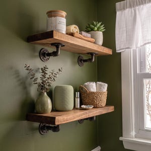 May include: Two horizontal wooden shelves mounted with black industrial pipe brackets. The top shelf holds a glass jar labeled Sea Salt Exfoliant, a loofah, and a succulent. The bottom shelf holds a vase with eucalyptus, a speaker, and a woven basket.