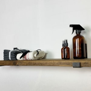 May include: A rustic wooden shelf with black metal brackets, holding folded gray and white towels, and amber glass spray bottles. The shelf is mounted on a white wall, creating a clean, minimalist aesthetic.