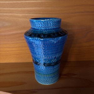 May include: A blue ceramic vase with a textured surface and a geometric pattern. The vase has a wide base and a narrow neck.
