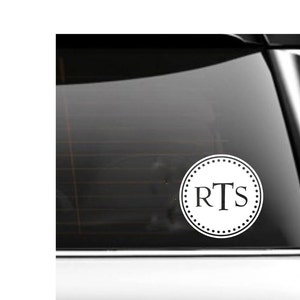 May include: White vinyl decal with a circle design and the letters "RTS" in the centre. The decal is on a car window.