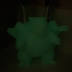 May include: A glowing green figurine of a creature with a rounded body, small legs, and arms. The figure is illuminated against a dark background, highlighting its form. The figurine appears to be made of a material that glows in the dark.