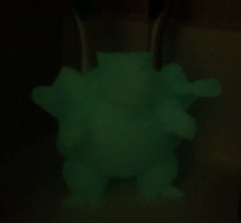 May include: A glowing green figurine of a creature with a rounded body, small legs, and arms. The figure is illuminated against a dark background, highlighting its form. The figurine appears to be made of a material that glows in the dark.