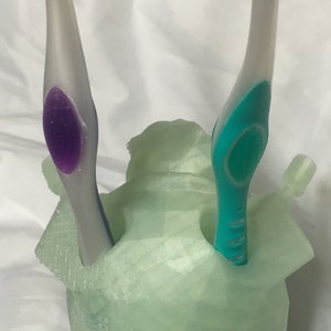 May include: A light green toothbrush holder with two toothbrushes. One toothbrush has a purple accent, and the other has a green accent. The holder is a unique, abstract shape.