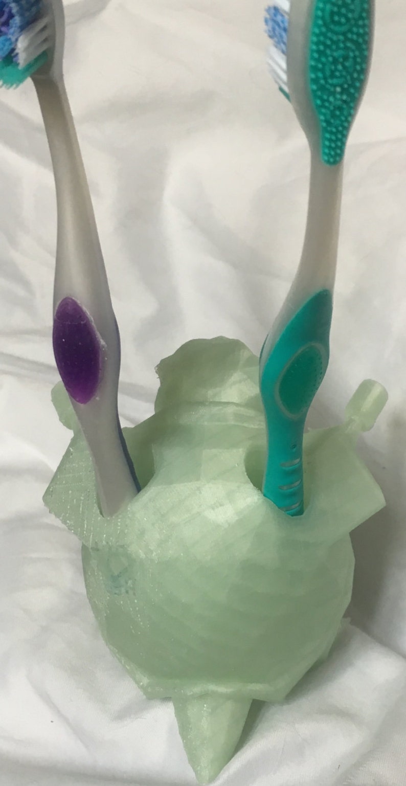 May include: A light green toothbrush holder with two toothbrushes. One toothbrush has a purple accent, and the other has a green accent. The holder is a unique, abstract shape.