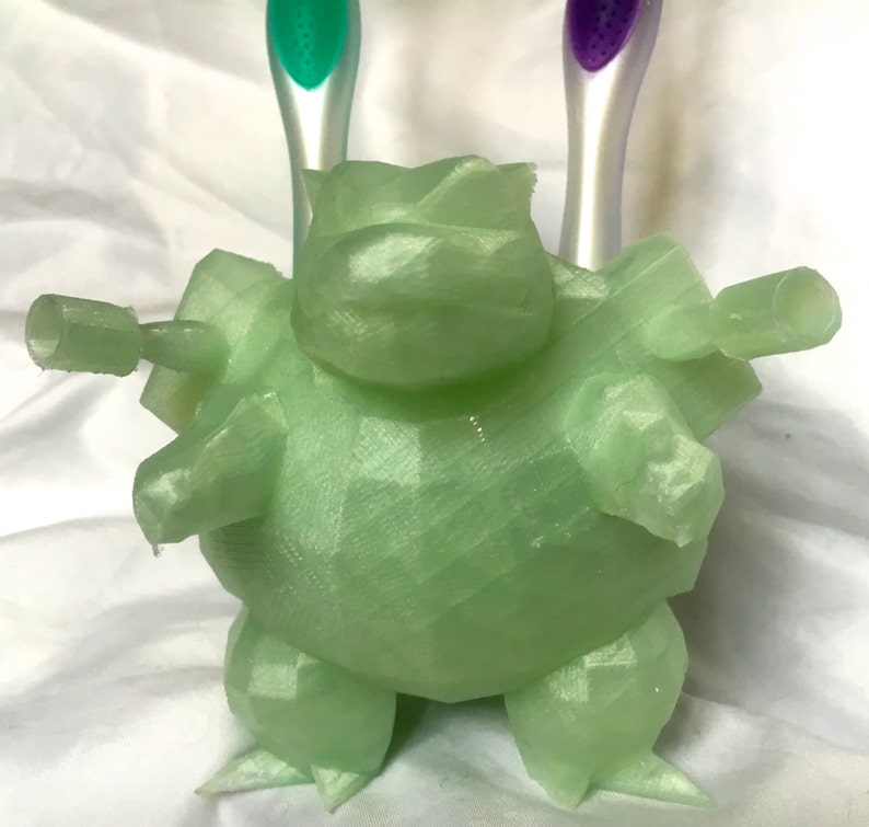 May include: A light green, 3D-printed toothbrush holder in the shape of a cartoon character. The holder has a geometric design and features two cylindrical arms to hold toothbrushes. Two toothbrushes with green and purple handles are visible.