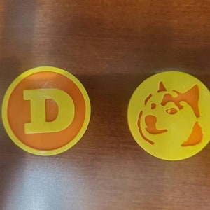May include: Two round, yellow and orange, 3D printed coasters. One coaster features the letter "D" in yellow. The other coaster features a yellow silhouette of a dog's head.
