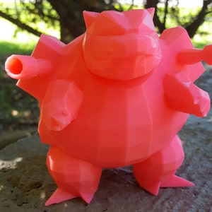 May include: A coral-colored, 3D-printed figure of a stylized creature with a geometric design. The figure has a rounded body, stubby legs, and two cylindrical protrusions on each side. The figure is standing on a stone surface, with a blurred green background.