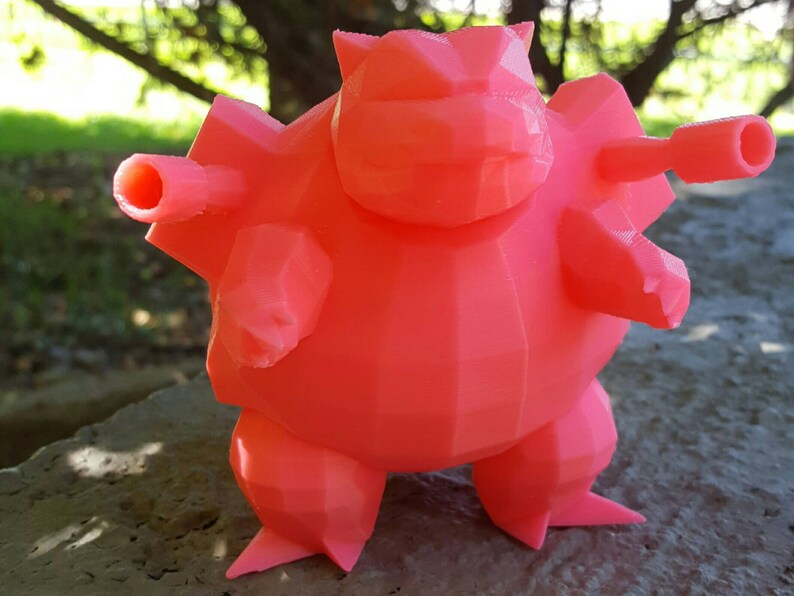 May include: A coral-colored, 3D-printed figure of a stylized creature with a geometric design. The figure has a rounded body, stubby legs, and two cylindrical protrusions on each side. The figure is standing on a stone surface, with a blurred green background.