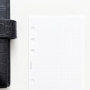 May include: A black leather planner with a crocodile pattern and a flap closure. A blank white gridded paper refill is next to the planner.
