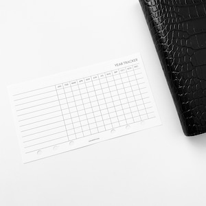 May include: A white year tracker printable with a grid of 12 columns representing each month of the year. The months are labeled January through December. The printable has six rows for tracking habits, goals, or other activities. The printable has six holes punched along the right side for use in a disc bound planner.