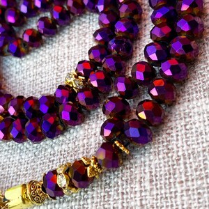 Op de afbeelding: Een ketting van paarse kristallen gebedskralen met gouden accenten. De kralen zijn gefacetteerd en hebben een glinsterende, iriserende afwerking.
