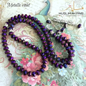Op de afbeelding: Een set gebedskralen, of tasbihs, gemaakt van metallic violet kristallen kralen. De kralen zijn geregen aan een zilveren ketting en hebben een zilveren bedel met de naam "NADINE" erop. De kralen worden getoond op een achtergrond met een bloemenpatroon.