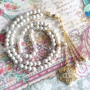 May include: A white pearl prayer beads necklace with a gold tassel and a gold pendant featuring a decorative design with clear rhinestones.