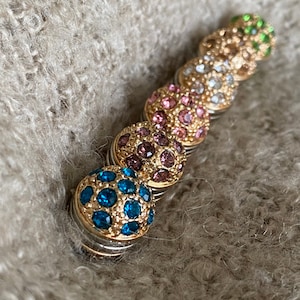 Könnte beinhalten: Ein Satz von sechs magnetischen Druckknöpfen mit goldfarbenen Metallfassungen und bunten Strasssteinen. Die Knöpfe sind in einer Reihe angeordnet, wobei der grüne Knopf oben und der blaue Knopf unten ist.