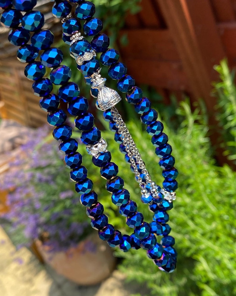 Op de afbeelding: Een blauwe gebedsketting met zilveren accenten. De kralen zijn gefacetteerd en hebben een metallic glans. De ketting is geregen aan een zilveren ketting en heeft een zilveren kwast.