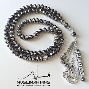 Op de afbeelding: Een zwarte kristallen gebedsketting mala met een zilveren bedel en een zilveren naamschild met de tekst "Rayyan".