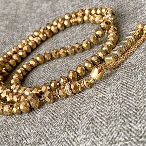 Op de afbeelding: Een gouden kralenketting met een gouden kwast en een bedel met de tekst "LAHIRA".