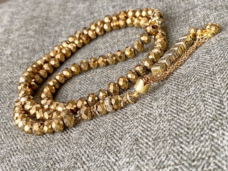 Op de afbeelding: Een gouden kralenketting met een gouden kwast en een bedel met de tekst "LAHIRA".