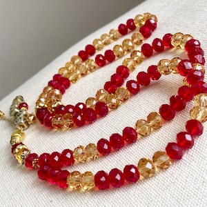 May include: A beaded necklace with alternating rows of red and gold-coloured faceted beads. The necklace has a decorative gold-coloured tassel and small cube-shaped beads with text. The beads are arranged in a layered design.