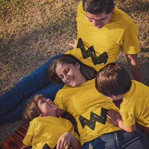 Peut inclure: Une famille de quatre personnes portant des t-shirts jaunes avec un motif en zigzag noir. Les t-shirts sont assortis et pr&eacute;sentent un personnage de dessin anim&eacute;. La famille est allong&eacute;e sur un tapis color&eacute; dans un espace herbeux.
