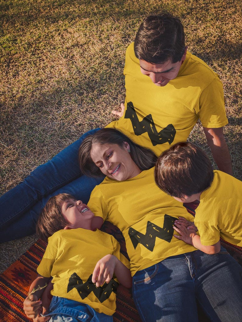Peut inclure: Une famille de quatre personnes portant des t-shirts jaunes avec un motif en zigzag noir. Les t-shirts sont assortis et pr&eacute;sentent un personnage de dessin anim&eacute;. La famille est allong&eacute;e sur un tapis color&eacute; dans un espace herbeux.